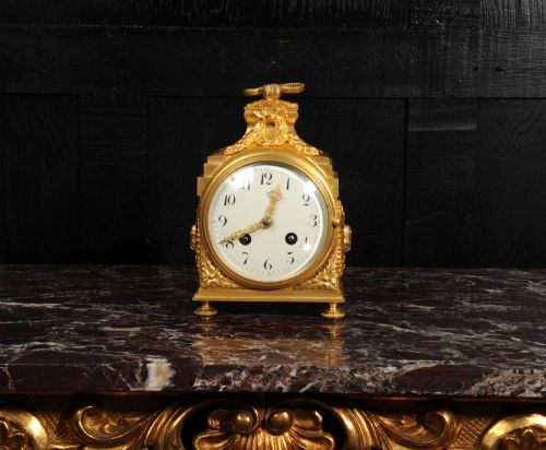 fig 1. An antique French gilt bronze ormolu officer clock with a ribbon top handle, decorative floral swags, and a white enamel dial with Arabic numerals.