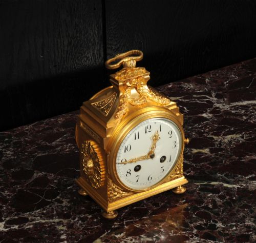 fig 2. An antique French gilt bronze ormolu officer clock with a ribbon top handle, decorative floral swags, and a white enamel dial with Arabic numerals.