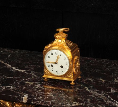 fig 3. An antique French gilt bronze ormolu officer clock with a ribbon top handle, decorative floral swags, and a white enamel dial with Arabic numerals.