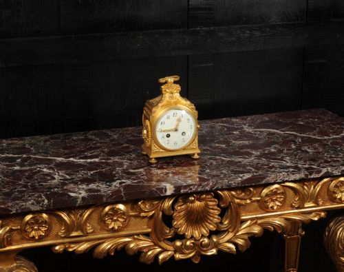 fig 4. An antique French gilt bronze ormolu officer clock with a ribbon top handle, decorative floral swags, and a white enamel dial with Arabic numerals.