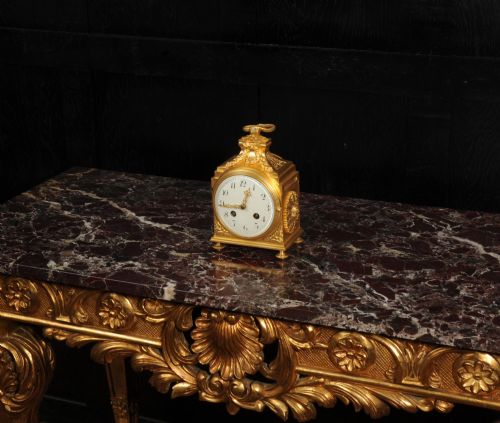 fig 5. An antique French gilt bronze ormolu officer clock with a ribbon top handle, decorative floral swags, and a white enamel dial with Arabic numerals.