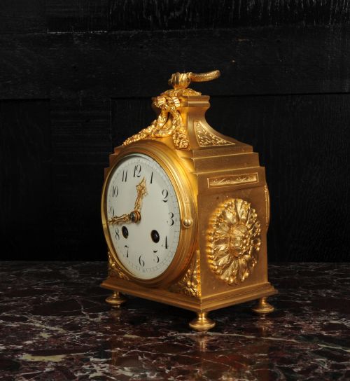 fig 7. An antique French gilt bronze ormolu officer clock with a ribbon top handle, decorative floral swags, and a white enamel dial with Arabic numerals.