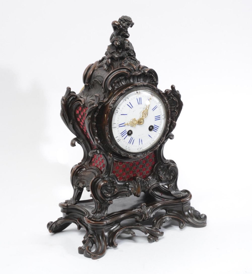An ornate dark bronze Rococo mantel clock featuring a cherub finial, scrolling rocaille design on a matching plinth, a white enamel dial with Roman numerals, and red textile backing panels.