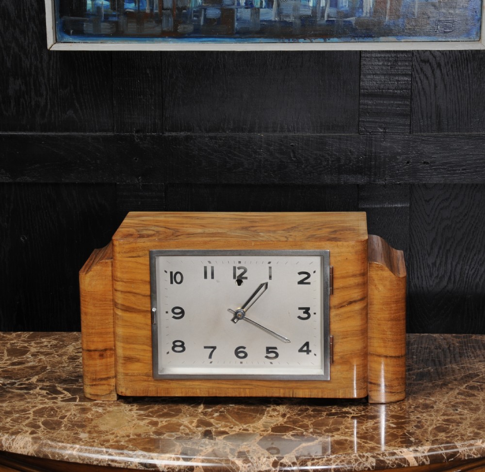 An Art Deco mantel clock featuring a figurative walnut veneered wooden case with stepped sides and a rectangular silver-toned dial with bold black numerals. View II