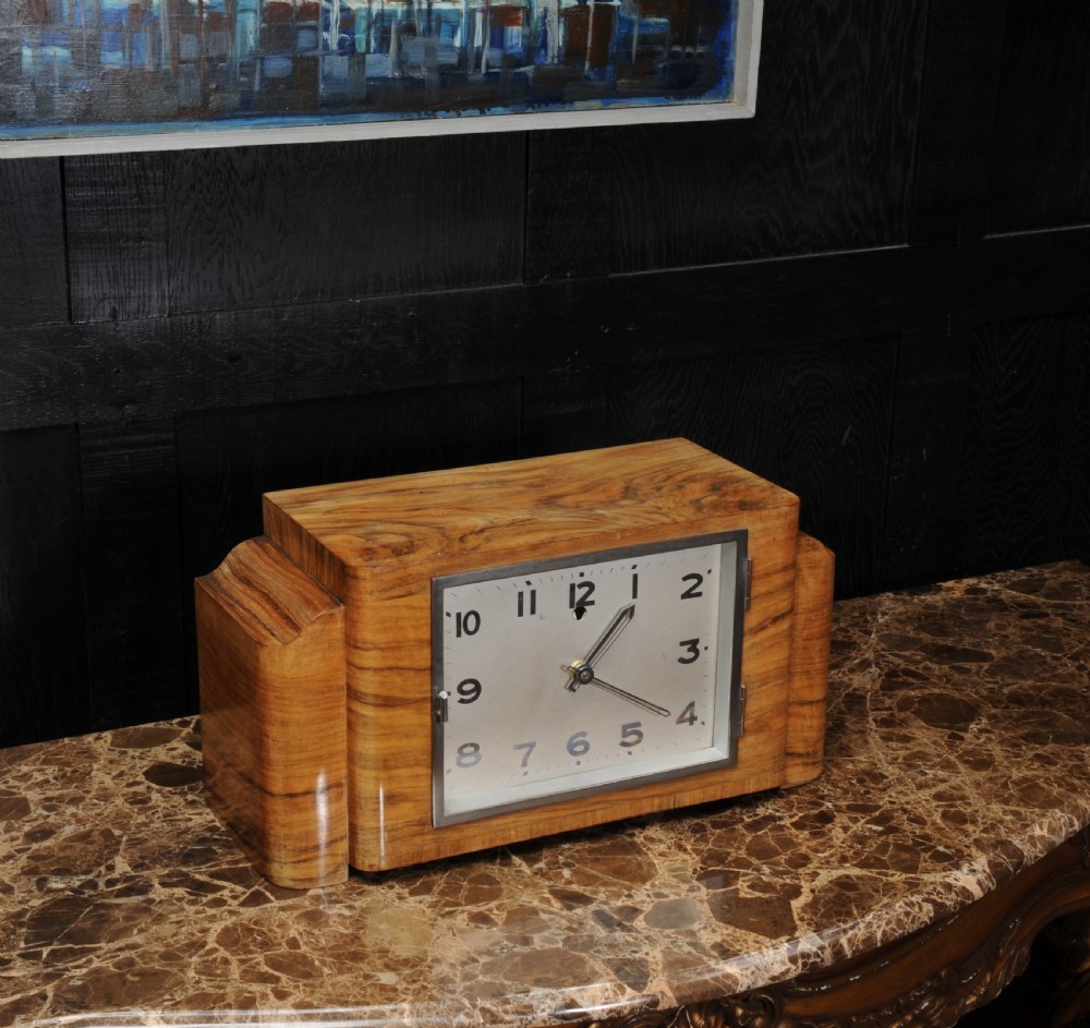 An Art Deco mantel clock featuring a figurative walnut veneered wooden case with stepped sides and a rectangular silver-toned dial with bold black numerals. View III