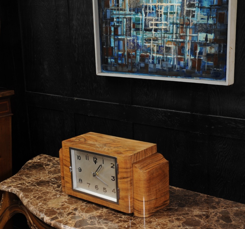 An Art Deco mantel clock featuring a figurative walnut veneered wooden case with stepped sides and a rectangular silver-toned dial with bold black numerals. View VI