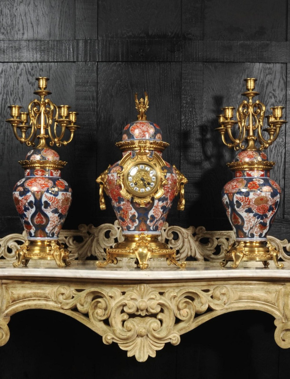 A three-piece antique French mantel garniture featuring a central Imari porcelain clock flanked by two matching gilt bronze candelabra atop porcelain urns.