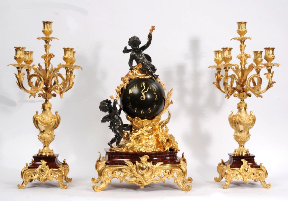An antique French Louis XV style clock garniture set featuring a central gilt bronze and dark bronze cherub clock on a red marble base, flanked by matching multi-arm gilt candelabra.
