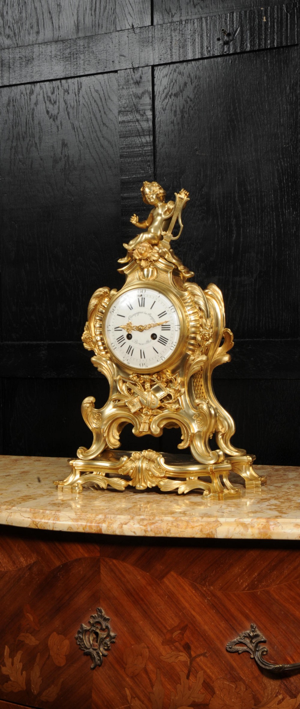 A French Rococo gilded bronze mantel clock featuring a cherub surmount, musical instrument reliefs, and a white enamel dial.