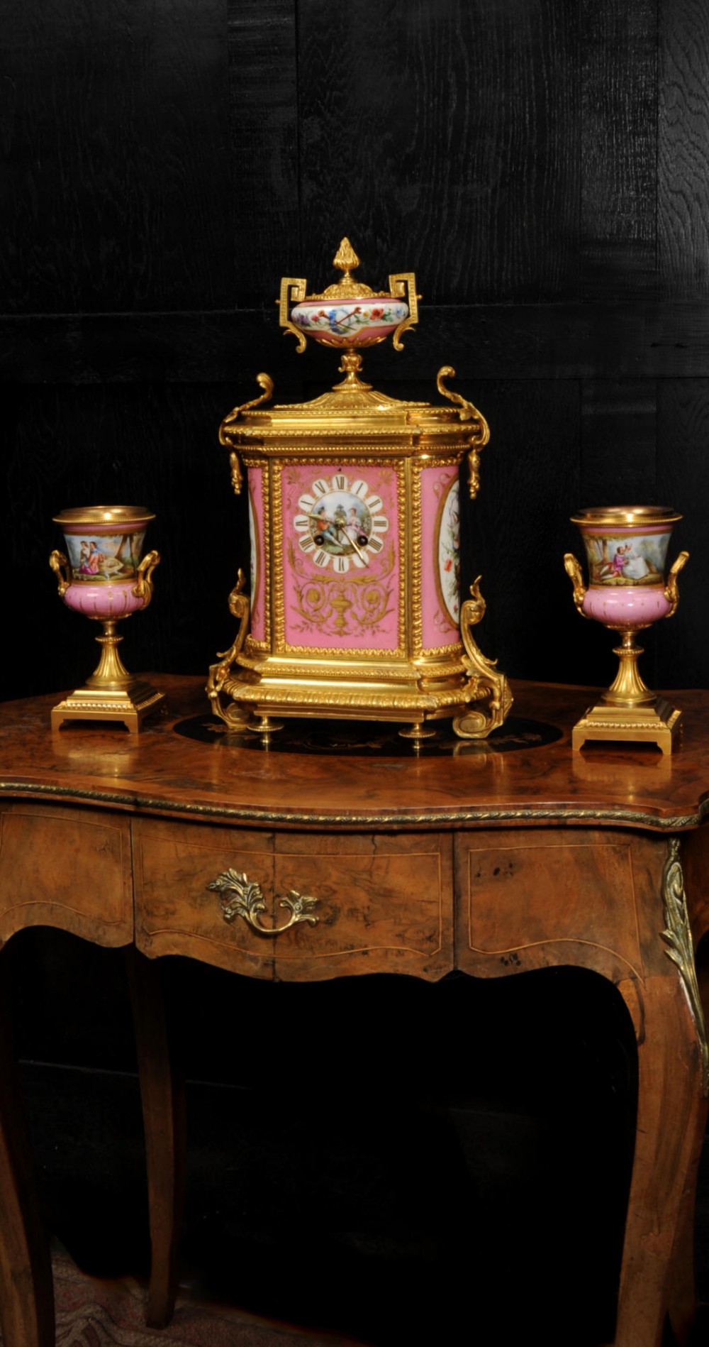 A French nineteenth-century clock garniture set comprising a gilt bronze and pink painted porcelain mantel clock accompanied by two matching ornamental urns.