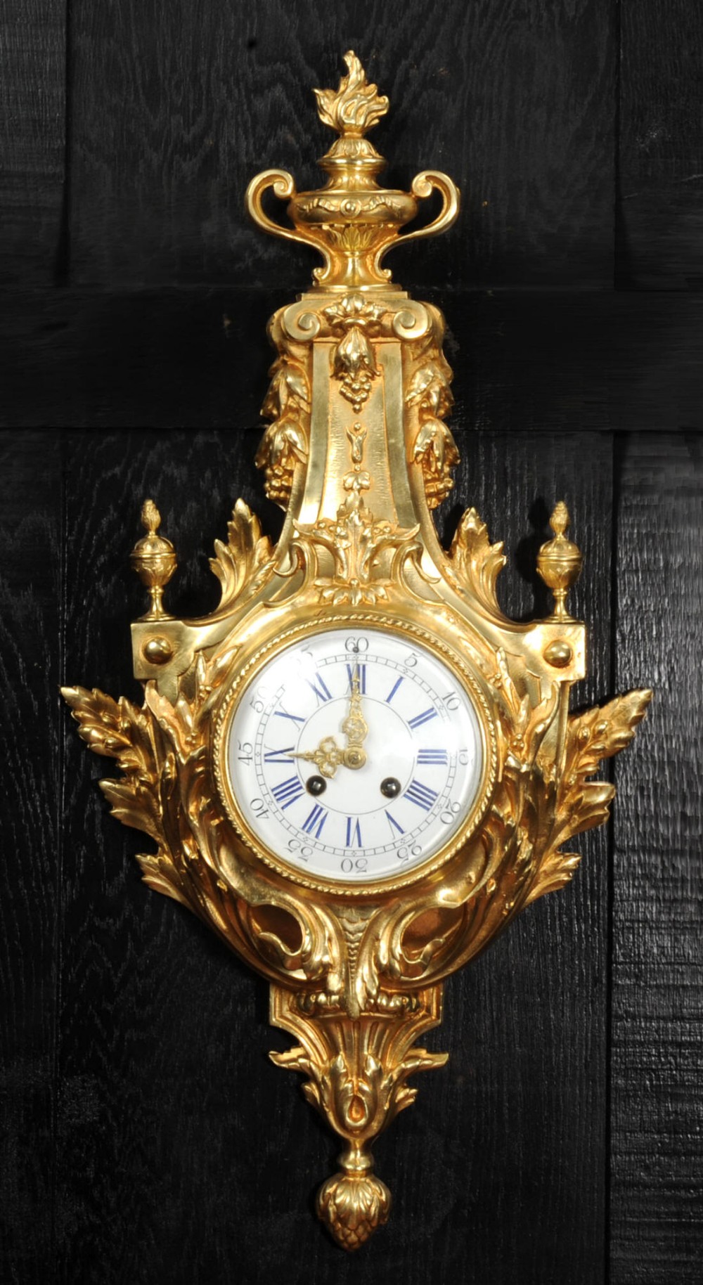 A Louis XVI style gilt bronze cartel wall clock featuring an urn finial, detailed acanthus leaf scrolls, and a white enamel dial with Roman numerals.