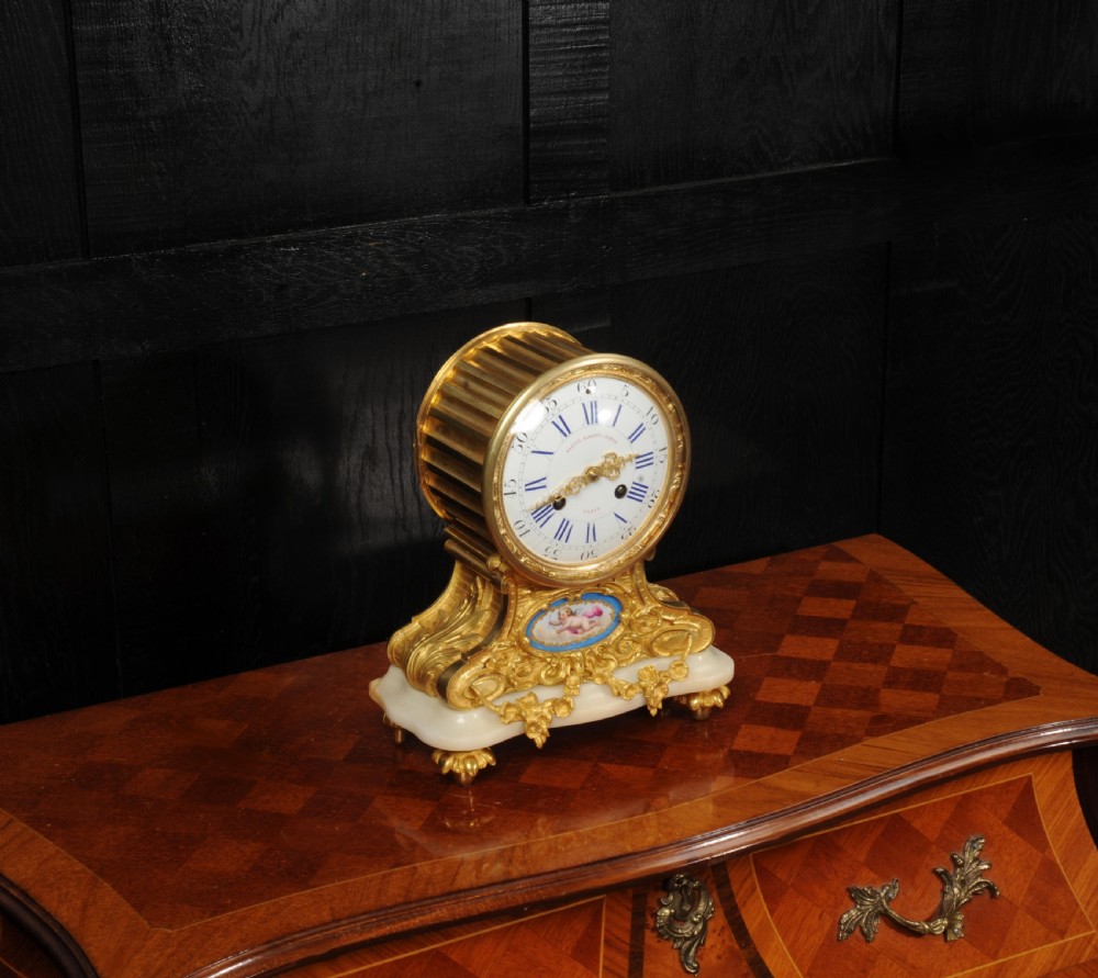 An antique French ormolu mantel clock featuring a white enamel dial, gilt bronze scrollwork casing, a painted cherub porcelain plaque, and a light marble base with bun feet. View VI