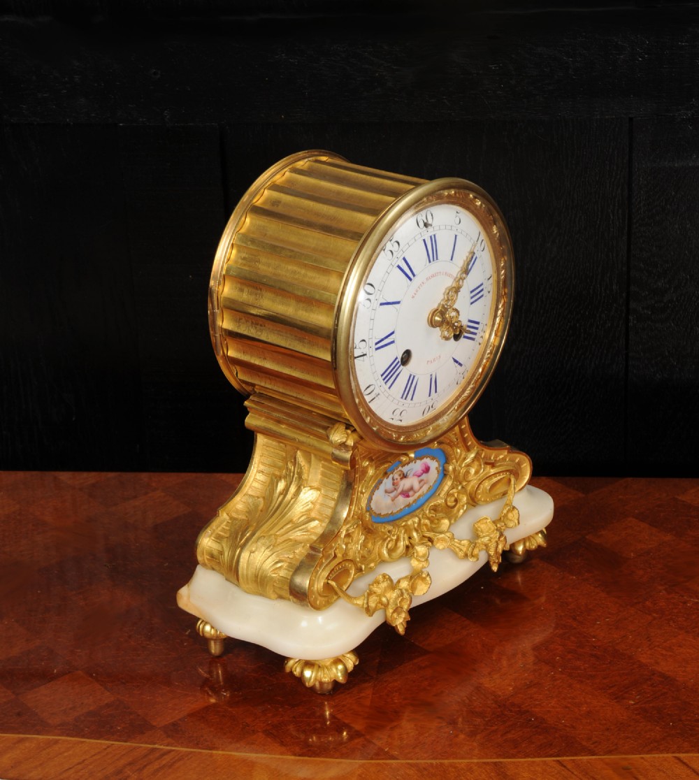 An antique French ormolu mantel clock featuring a white enamel dial, gilt bronze scrollwork casing, a painted cherub porcelain plaque, and a light marble base with bun feet. View IX