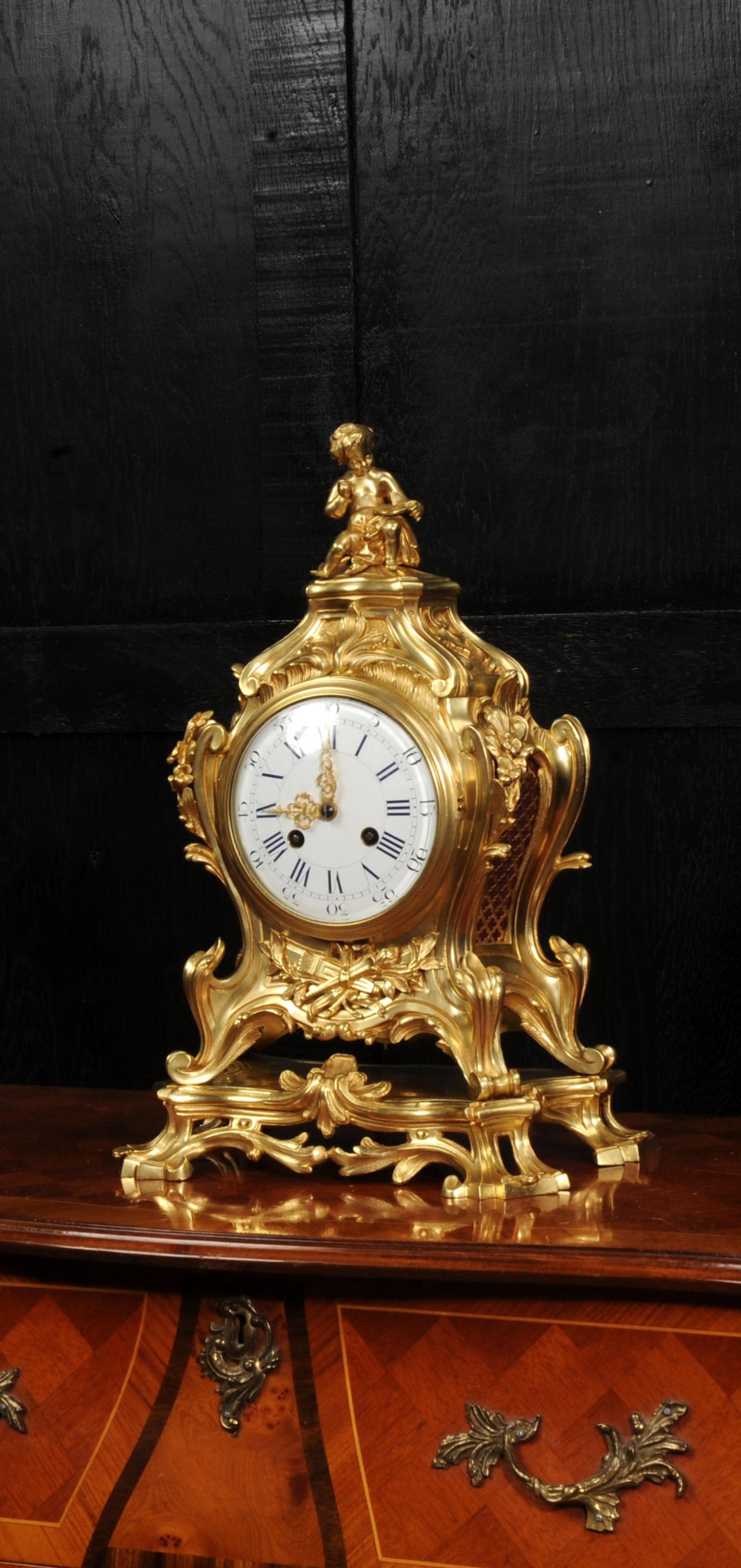 An antique French gilt bronze Rococo mantel clock featuring an ornate scrollwork case topped with a seated cherub figure and a white enamel dial.