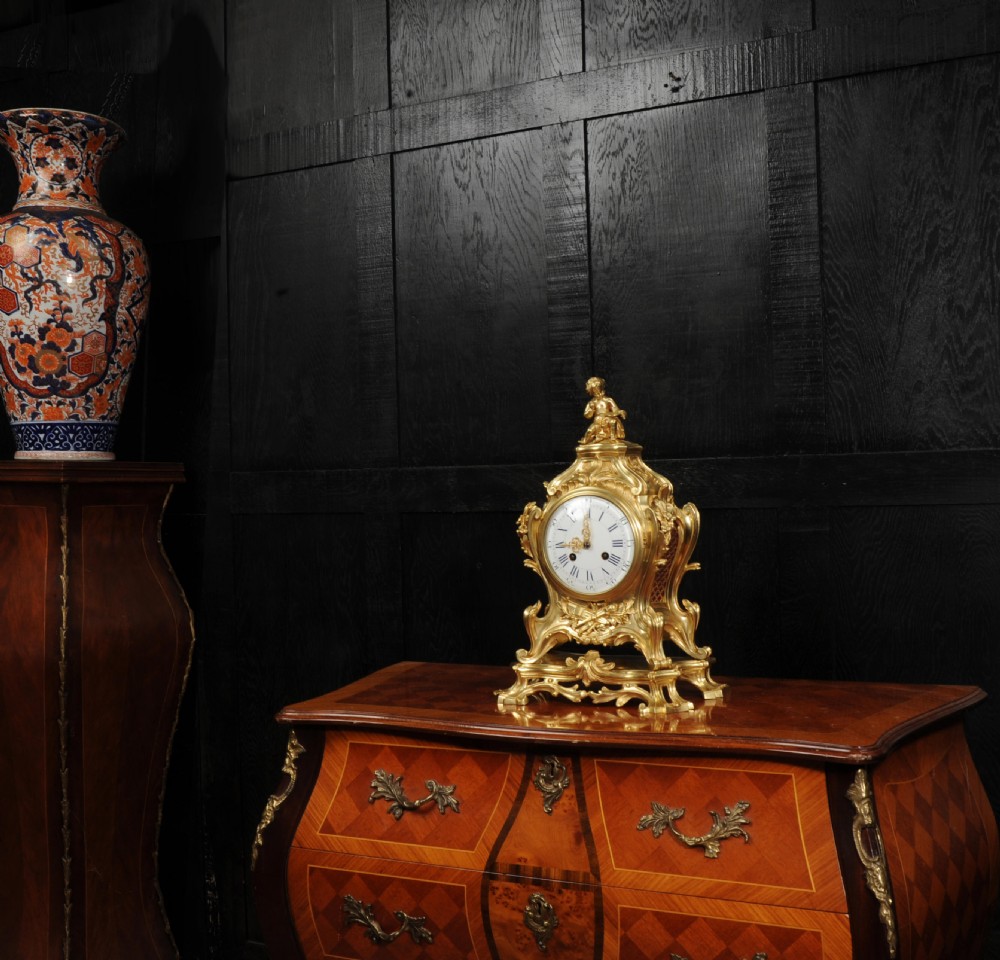 An antique French gilt bronze Rococo mantel clock featuring an ornate scrollwork case topped with a seated cherub figure and a white enamel dial. View V