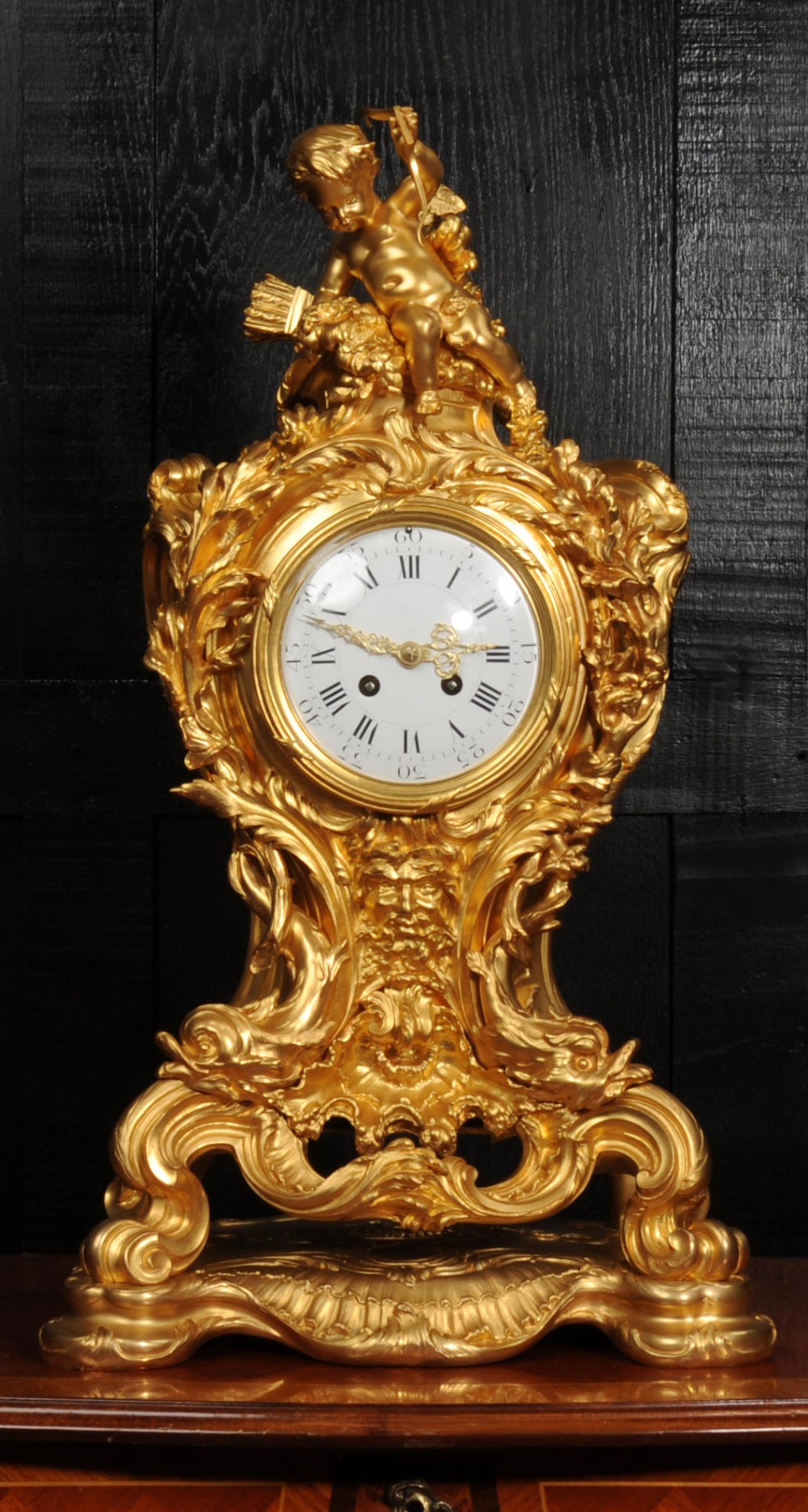 An ornate French Rococo ormolu mantel clock decorated with an elaborate cherub finial, acanthus leaf scrolls, and a white enamel dial.