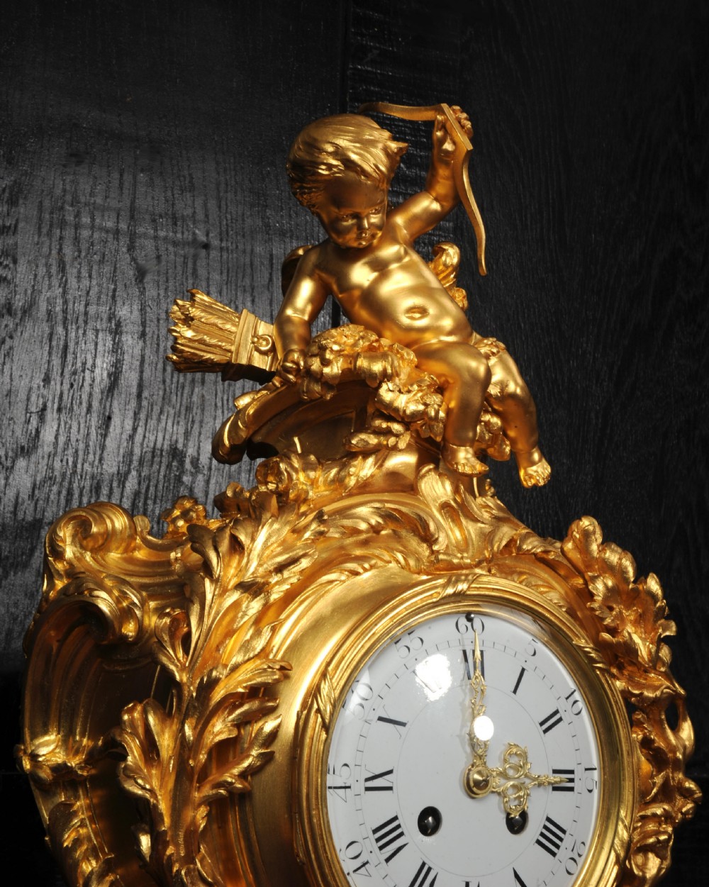 An ornate French Rococo ormolu mantel clock decorated with an elaborate cherub finial, acanthus leaf scrolls, and a white enamel dial. View V
