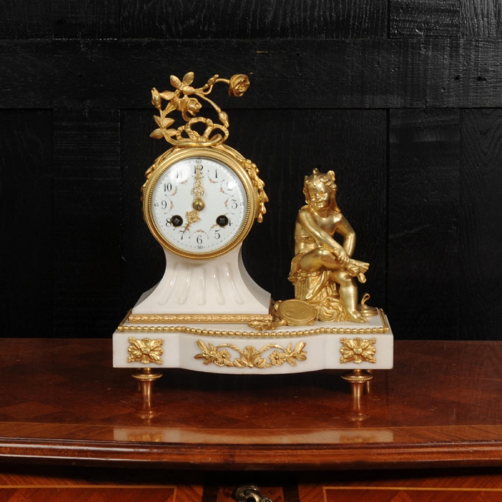 An antique French mantel clock featuring a white marble base decorated with gilt bronze mounts, an off-centre enamel dial, and a gilded bronze putto figure seated alongside. View II