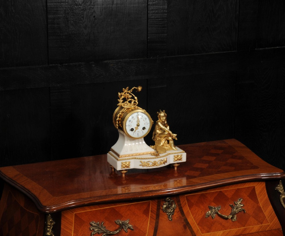 An antique French mantel clock featuring a white marble base decorated with gilt bronze mounts, an off-centre enamel dial, and a gilded bronze putto figure seated alongside. View III
