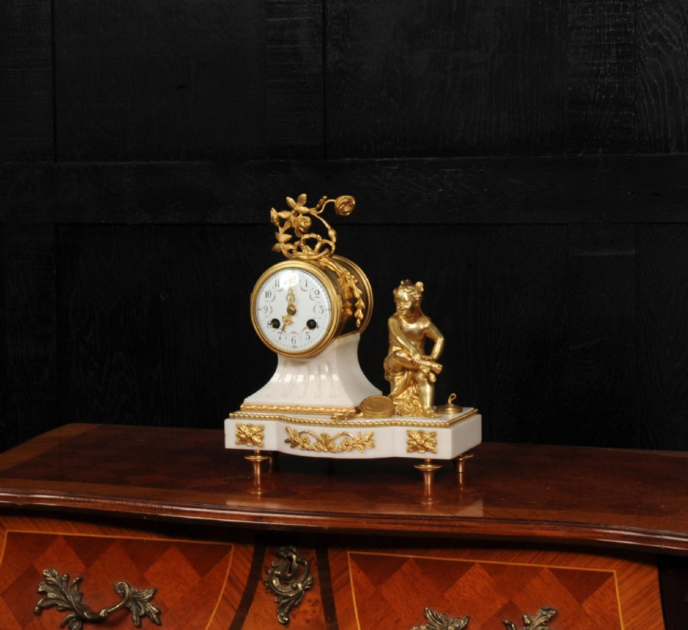 An antique French mantel clock featuring a white marble base decorated with gilt bronze mounts, an off-centre enamel dial, and a gilded bronze putto figure seated alongside. View IV