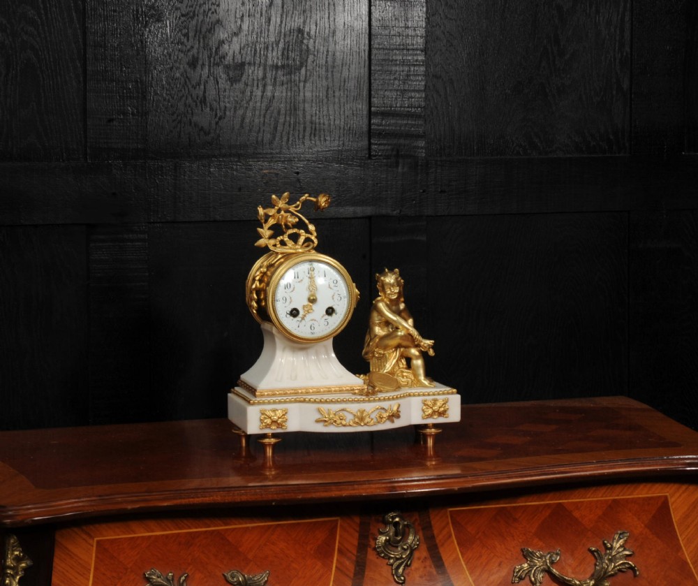 An antique French mantel clock featuring a white marble base decorated with gilt bronze mounts, an off-centre enamel dial, and a gilded bronze putto figure seated alongside. View V