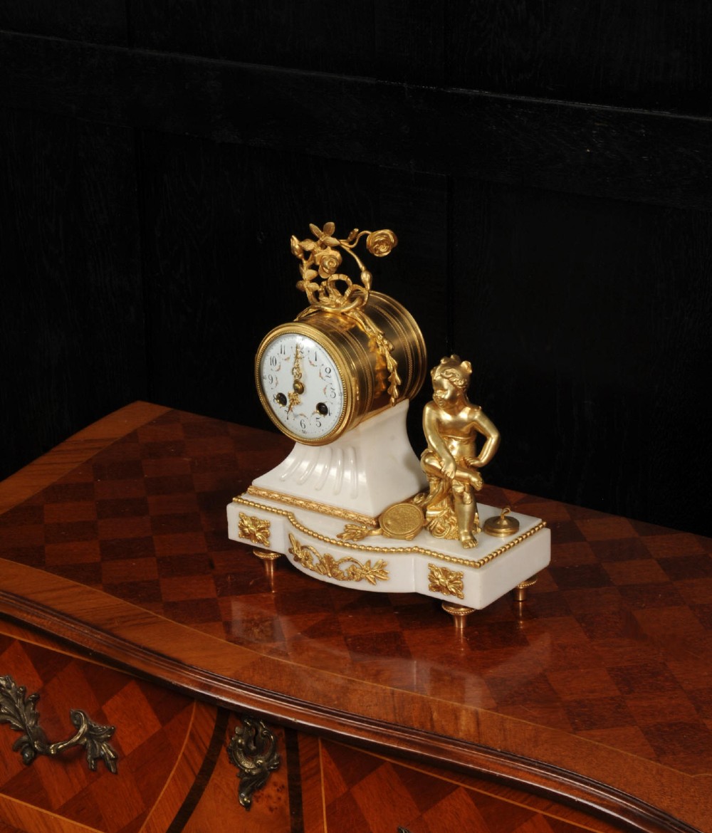 An antique French mantel clock featuring a white marble base decorated with gilt bronze mounts, an off-centre enamel dial, and a gilded bronze putto figure seated alongside. View VII