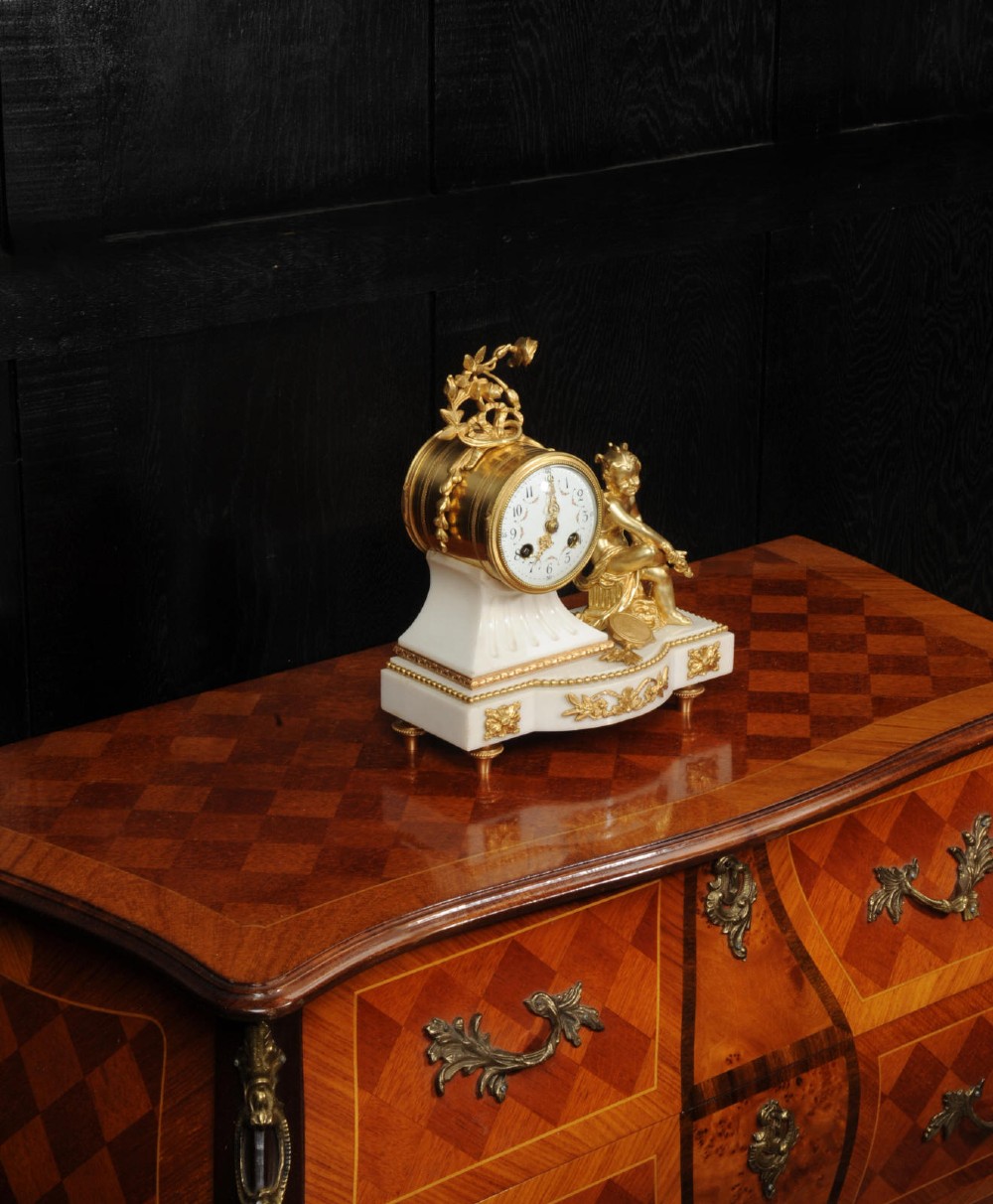 An antique French mantel clock featuring a white marble base decorated with gilt bronze mounts, an off-centre enamel dial, and a gilded bronze putto figure seated alongside. View VIII