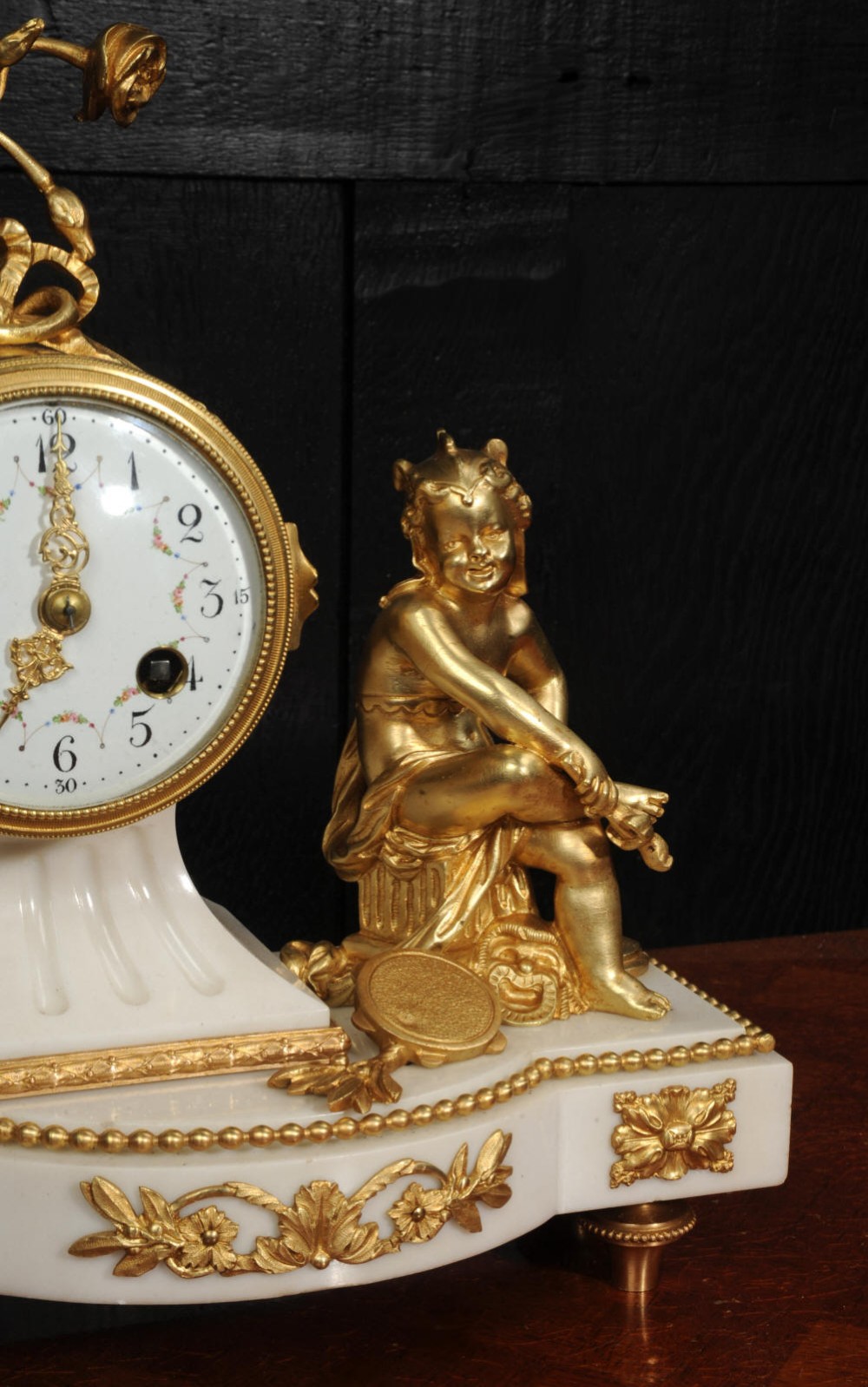 An antique French mantel clock featuring a white marble base decorated with gilt bronze mounts, an off-centre enamel dial, and a gilded bronze putto figure seated alongside. View IX