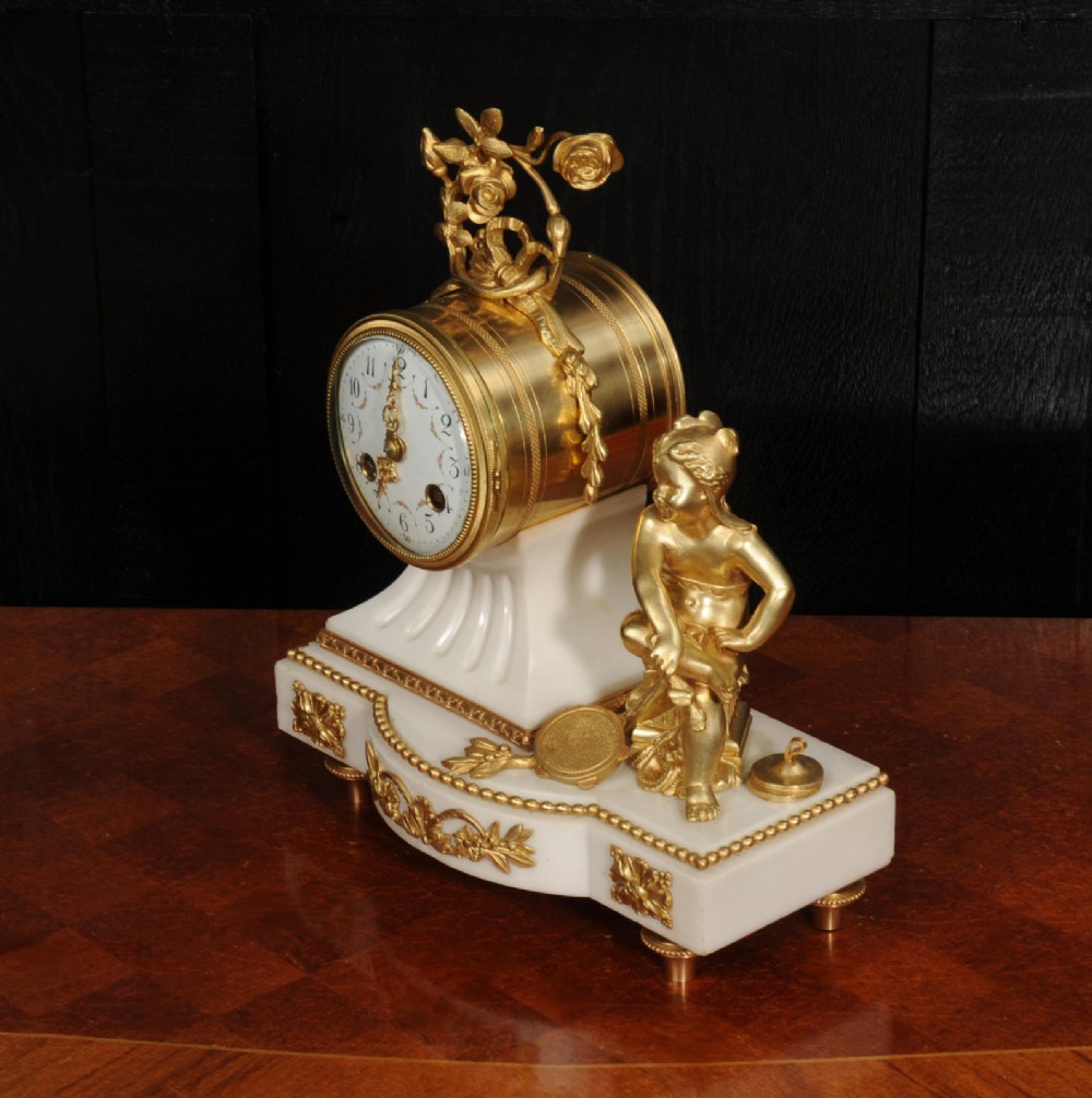 An antique French mantel clock featuring a white marble base decorated with gilt bronze mounts, an off-centre enamel dial, and a gilded bronze putto figure seated alongside. View X