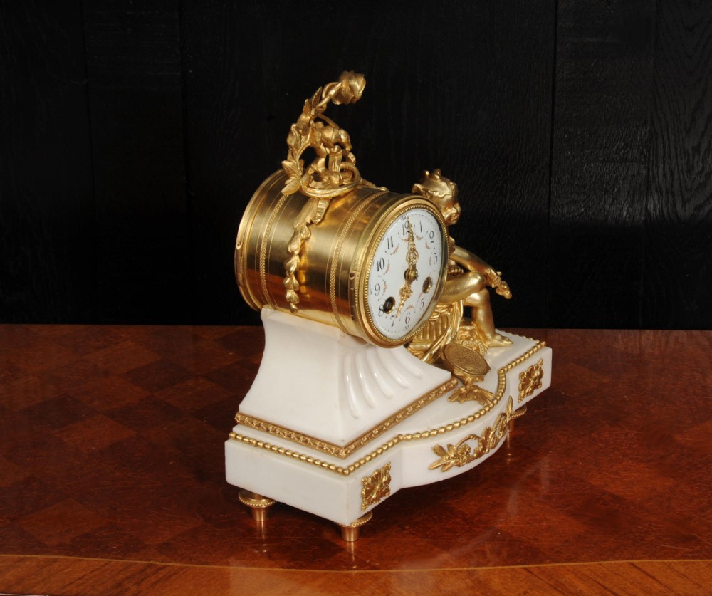 An antique French mantel clock featuring a white marble base decorated with gilt bronze mounts, an off-centre enamel dial, and a gilded bronze putto figure seated alongside. View XI