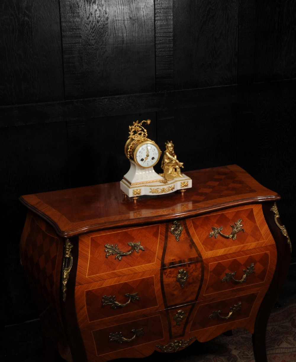 An antique French mantel clock featuring a white marble base decorated with gilt bronze mounts, an off-centre enamel dial, and a gilded bronze putto figure seated alongside. View XII