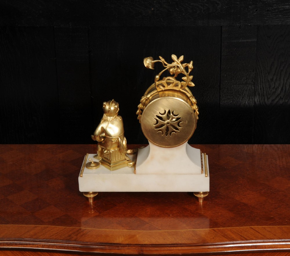 An antique French mantel clock featuring a white marble base decorated with gilt bronze mounts, an off-centre enamel dial, and a gilded bronze putto figure seated alongside. View XIII