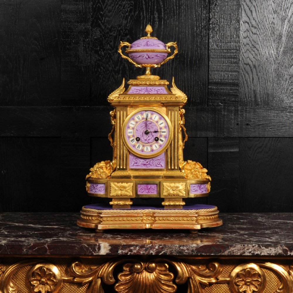 An ornate French gilt bronze and lilac hand-painted porcelain mantel clock featuring a decorative urn finial and fluted side columns.