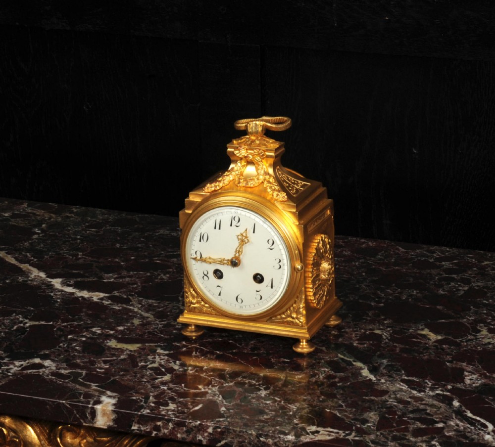 An antique French gilt bronze ormolu officer clock with a ribbon top handle, decorative floral swags, and a white enamel dial with Arabic numerals. View IV