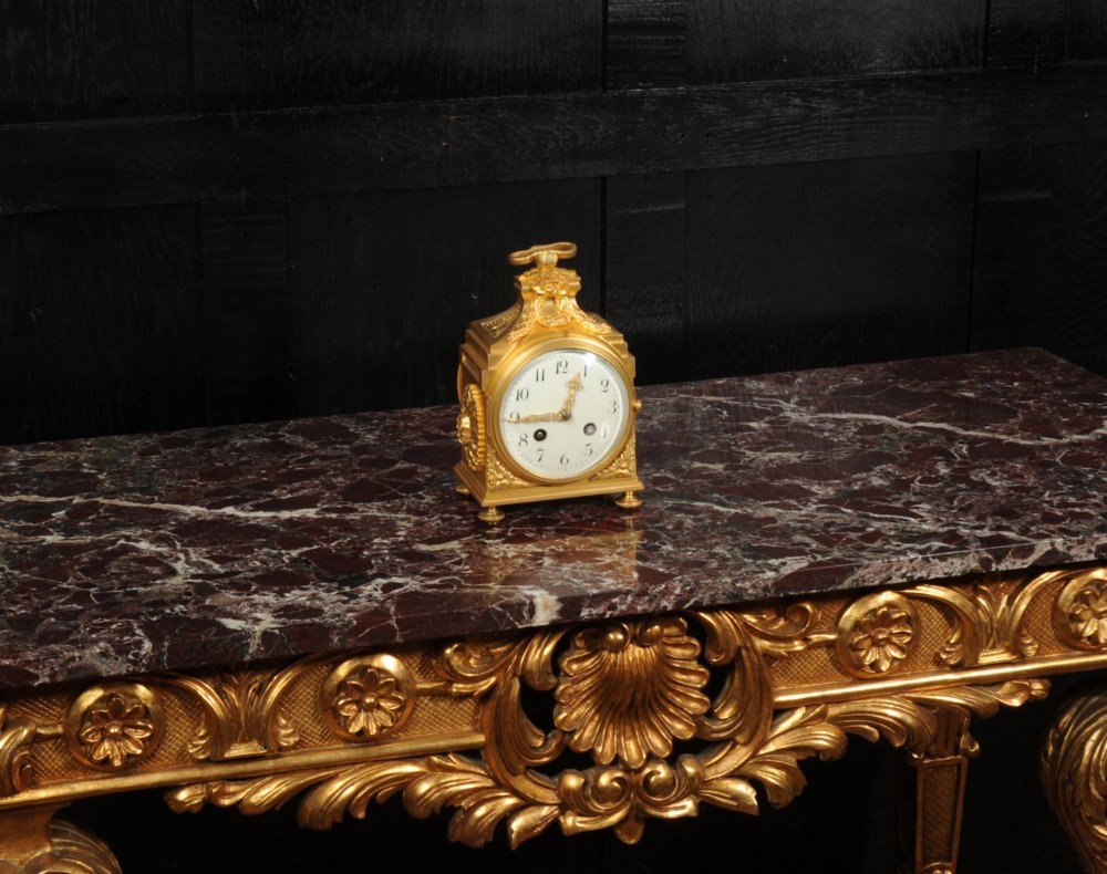 An antique French gilt bronze ormolu officer clock with a ribbon top handle, decorative floral swags, and a white enamel dial with Arabic numerals. View V
