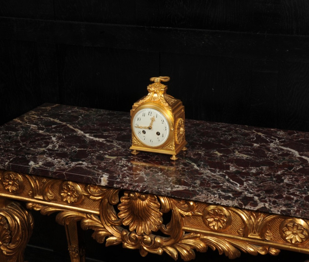 An antique French gilt bronze ormolu officer clock with a ribbon top handle, decorative floral swags, and a white enamel dial with Arabic numerals. View VI