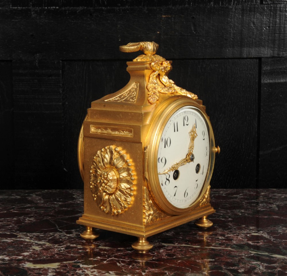 An antique French gilt bronze ormolu officer clock with a ribbon top handle, decorative floral swags, and a white enamel dial with Arabic numerals. View VII