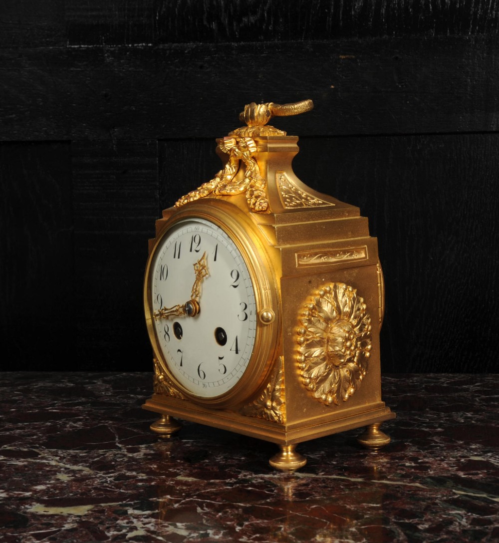 An antique French gilt bronze ormolu officer clock with a ribbon top handle, decorative floral swags, and a white enamel dial with Arabic numerals. View VIII