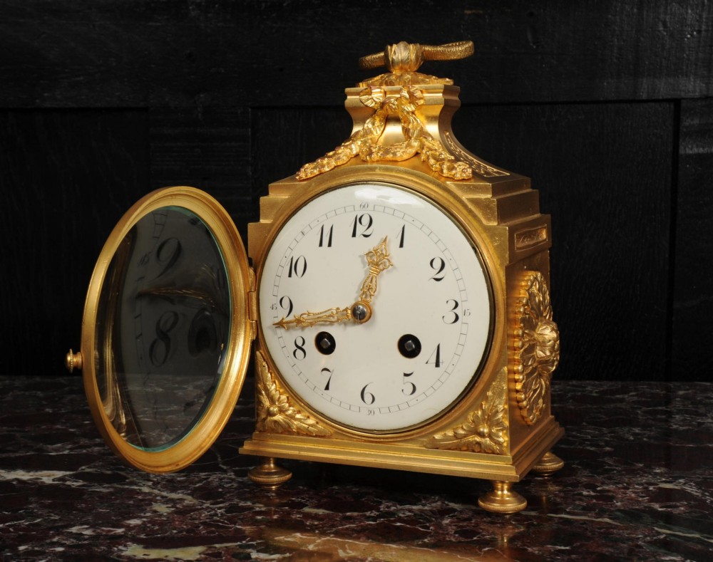 An antique French gilt bronze ormolu officer clock with a ribbon top handle, decorative floral swags, and a white enamel dial with Arabic numerals. View X