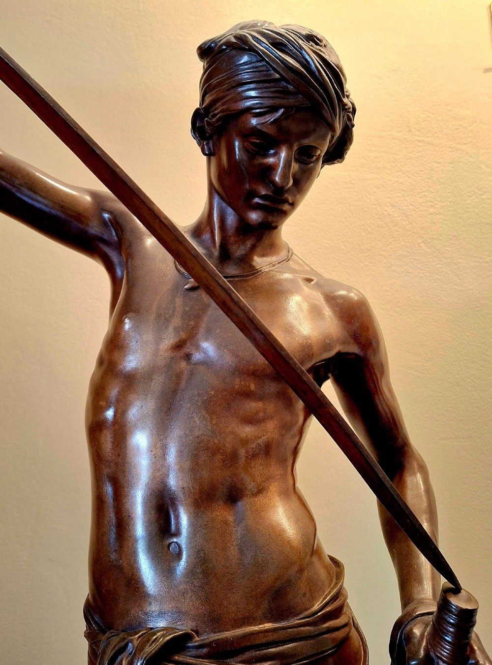 A patinated bronze sculpture of a youth holding a sword with one foot resting on a severed head, mounted on a circular base with an inscribed plinth. View XI