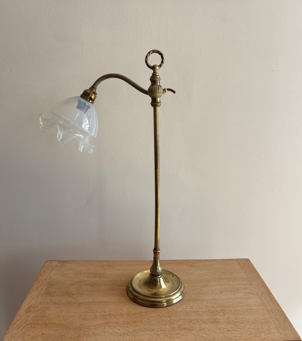 An Edwardian adjustable brass desk lamp featuring a circular stepped base, a vertical pole with a ring finial, a curved arm, and a translucent frilled glass shade.