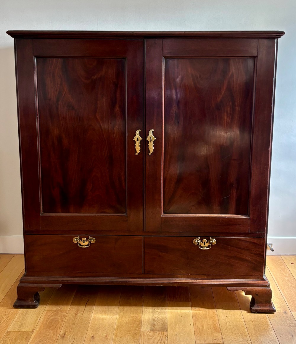 A dark George II mahogany press cupboard featuring two large panelled doors with brass escutcheons above two drawers on bracket feet.