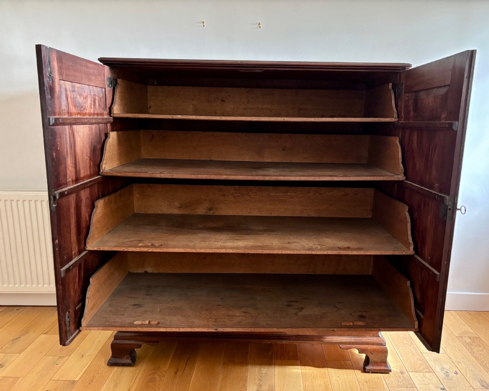 A dark George II mahogany press cupboard featuring two large panelled doors with brass escutcheons above two drawers on bracket feet. View II