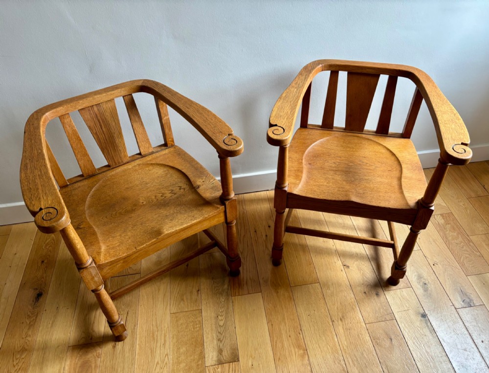 A pair of solid oak Edwardian armchairs featuring curved top rails, carved scroll handrests, vertical back splats, and contoured saddle seats.