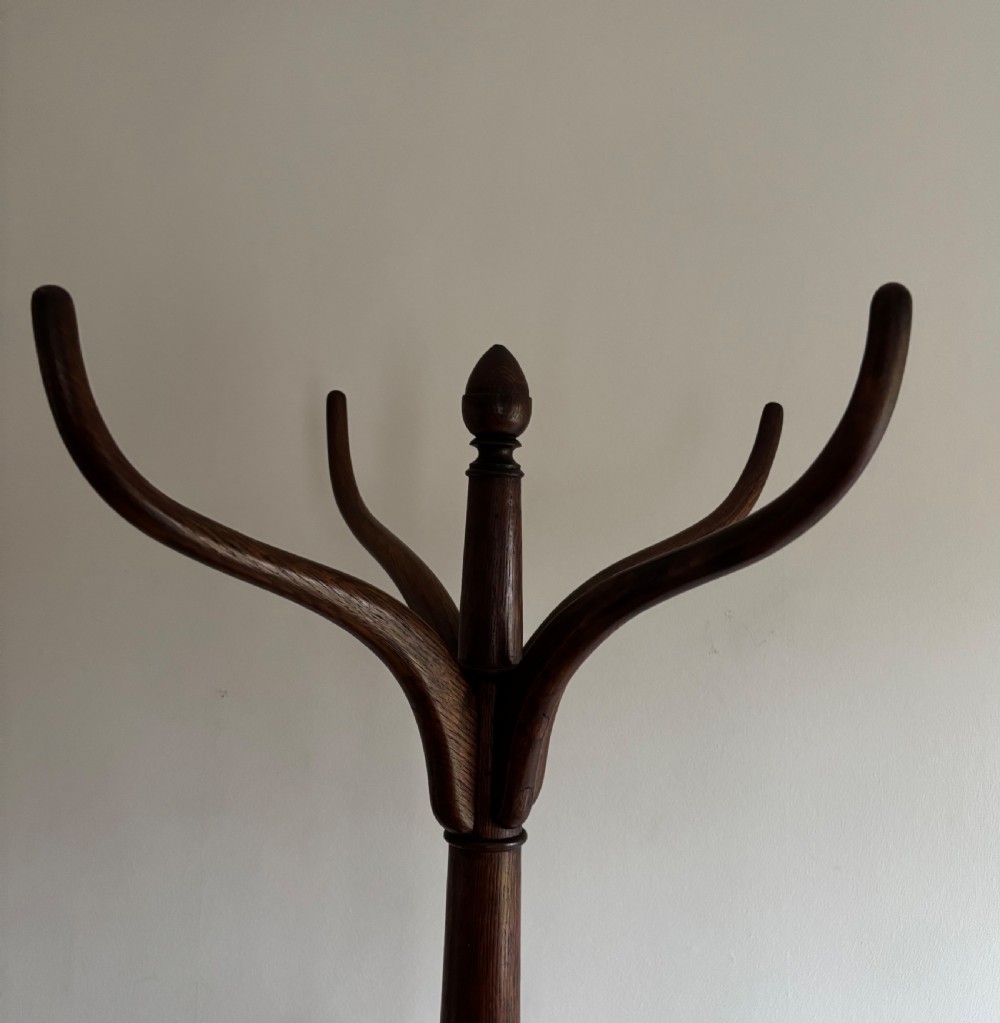 A dark oak Regency coat stand featuring multiple carved hooks, a central turned post, a circular brass umbrella ring, and splayed legs over a rounded drip tray. View II
