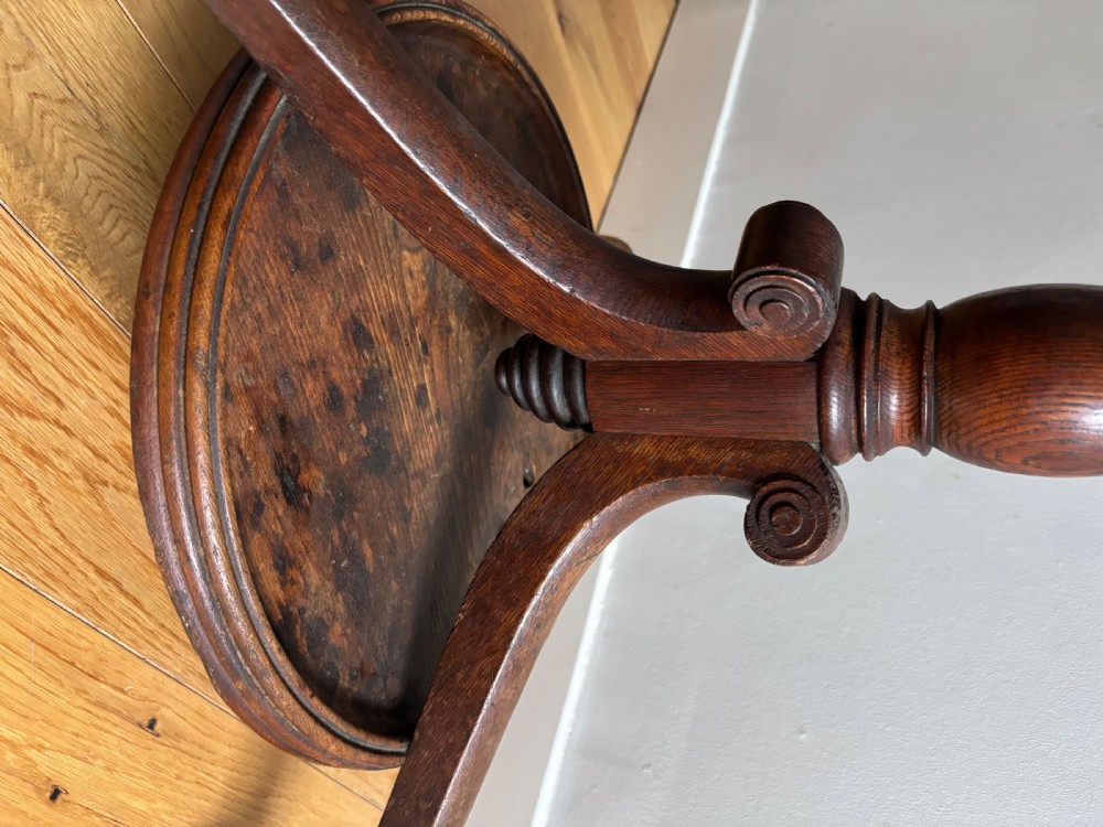 A dark oak Regency coat stand featuring multiple carved hooks, a central turned post, a circular brass umbrella ring, and splayed legs over a rounded drip tray. View VI