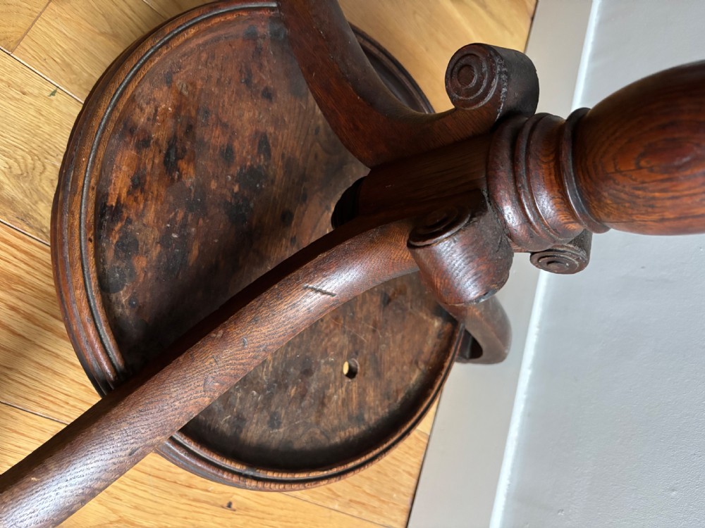 A dark oak Regency coat stand featuring multiple carved hooks, a central turned post, a circular brass umbrella ring, and splayed legs over a rounded drip tray. View VII