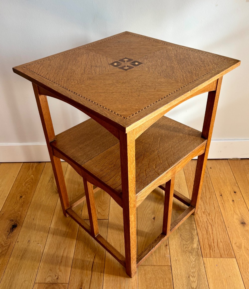 An antique Arts and Crafts square oak occasional table featuring a central marquetry motif, banded inlay border, lower tier shelf, and straight legs.