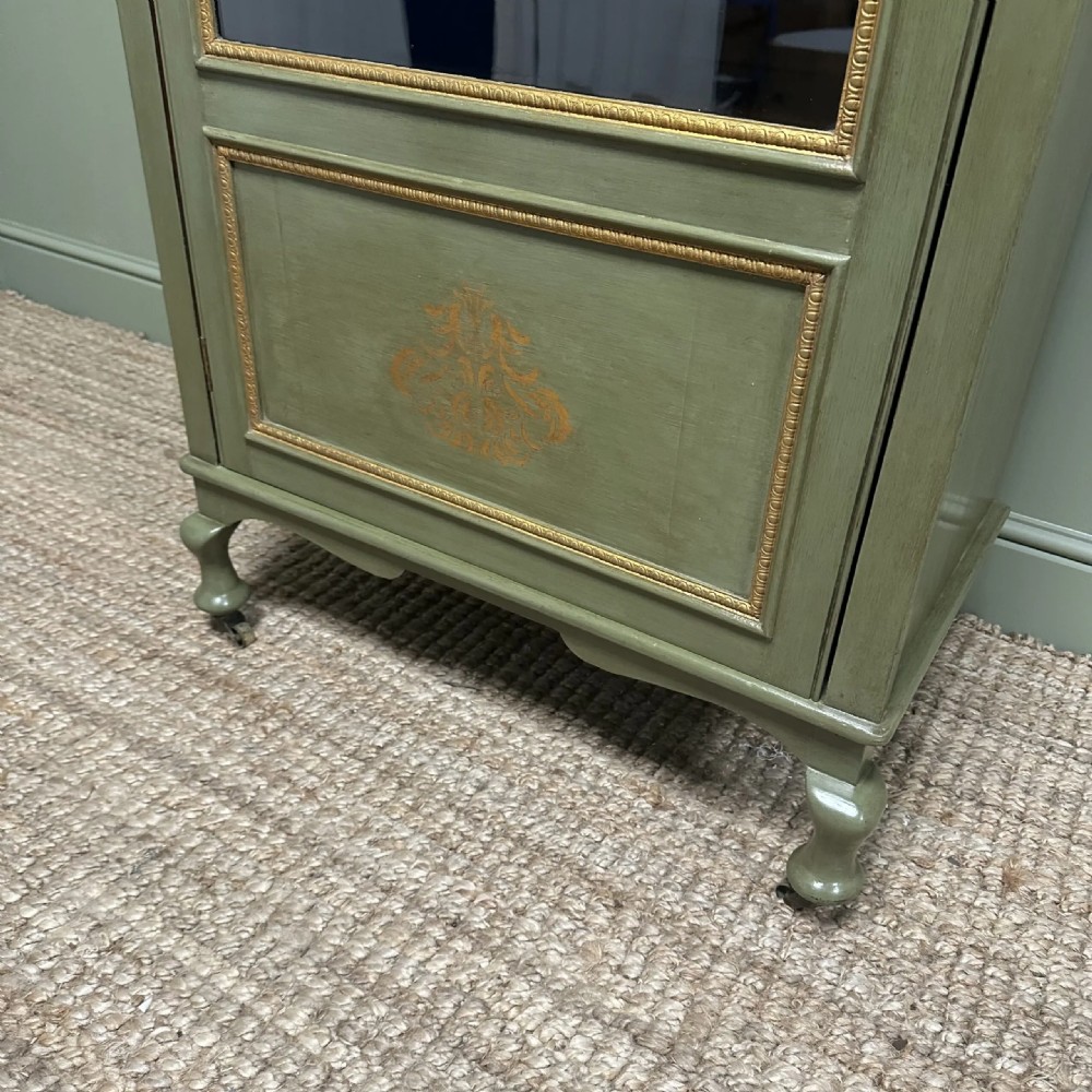 A muted green Edwardian display cabinet featuring a shaped pediment with gilt ornamentation, a single glazed door, and cabriole legs. View IV
