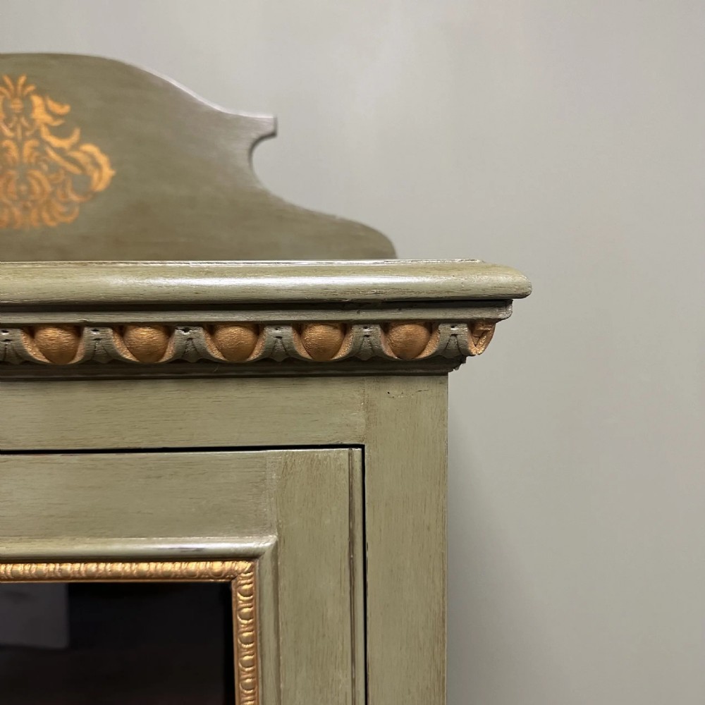 A muted green Edwardian display cabinet featuring a shaped pediment with gilt ornamentation, a single glazed door, and cabriole legs. View VI
